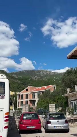 A daytime video shows a mountainous landscape with two distinct buildings. In the foreground, partially constructed, is a building with exposed brickwork and a red-tiled roof. It features several windows and a large circular opening on one side. Beyond this, a lush green, tree-covered slope leads up to a rocky mountain. Perched on a prominent, rocky outcrop higher on the mountain is a finished, white building with large windows and balconies, suggesting a modern residence or resort. The sky is bright blue with scattered white clouds. The camera initially captures a wider view, then zooms in to focus on the building on the mountain, and finally zooms back out to the original wider perspective. The audio includes ambient sounds, likely from passing vehicles or the environment. The scene is set in Vrijesno, Montenegro. No people or visible text are present.