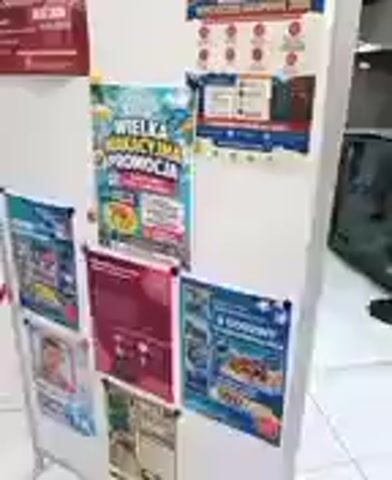 The video shows a close-up view of a bulletin board displaying various posters. The posters appear to be advertisements or promotional materials. The bulletin board is white and rectangular, mounted on a silver metal stand. There are several rows of posters, with some overlap and slight distortions due to the camera angle and focus.

The posters are colorful and varied in their content. One prominent poster in the center has large, bold text in what appears to be Polish, featuring tropical imagery and text like "WIELKA WAKACYJNA PROMOCJA" which translates to "BIG VACATION PROMOTION." Another poster to its right has a grid of circular images and text that is difficult to read. Below these, more posters are visible, including one with what looks like a person's face and another with images of food. The overall impression is that of a commercial setting, possibly a store, a travel agency, or a community center, where these promotions are displayed.

The lighting suggests an indoor environment, possibly during daytime given the brightness, but no specific time of day can be determined. The background is slightly out of focus, showing a glimpse of what might be electronic equipment or office furniture. The scene is static, with no discernible activity other than the implied purpose of displaying information. The focus is solely on the bulletin board and its contents. The location context of Reda, Poland, might suggest that the text on the posters is in Polish, which is consistent with the visible phrases.