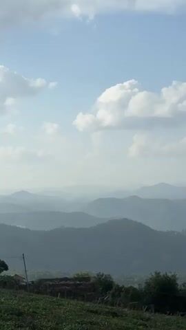 The video shows a landscape of rolling hills under a hazy sky with scattered clouds. The foreground features greenery, possibly a field or vegetation, with a wooden post and wire on the left. The hills recede into the distance, layered in shades of blue and gray due to atmospheric perspective. The overall impression is one of a vast, serene natural environment.