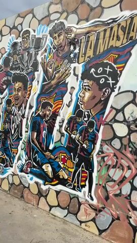 The video shows a mural of soccer players on a textured wall. The mural features several depictions of players, including one large portrait on the left, and smaller figures in action and celebrating with a trophy. The wall itself has a background of irregular shapes resembling stones in various colors. To the left of the mural, there is a concrete walkway and a small soccer goal with two soccer balls on the ground. Further down the walkway, the wall continues with more graffiti, including large, colorful lettering. The camera then pans to show a wider view of an outdoor area with a concrete ground, trees, buildings, and a clear sky with some clouds. In the distance, two young boys are visible on the concrete expanse, with one boy appearing to be running and the other walking. A triangular structure with birds perched on it is also visible in the background. The sun is shining brightly, casting shadows.