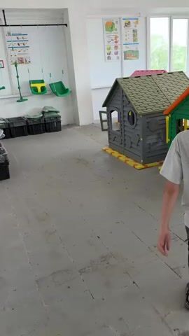 The video shows the interior of a large, brightly lit room that appears to be a children's playroom or activity center. The camera starts by showing several small, colorful playhouses lined up against a wall. A child wearing a gray shirt and dark shorts walks past the houses on the right. The camera then pans to the right, passing behind a white pillar, to reveal more of the room. 

The room contains several tables and chairs, with some tables covered in large, colorful building blocks. There are multiple sets of colorful, geometrically-shaped tables and chairs. Large windows line the far wall, offering a view of buildings and trees outside. There are posters hanging on the walls, likely with educational or safety-related content for children. In one area, there is a table covered with a large collection of blue and red building blocks. Overall, the space looks like it is set up for children to play and learn, but it appears a bit sparse or unfinished.
