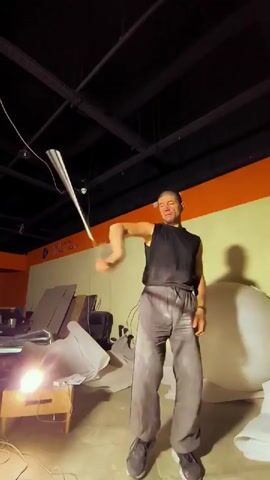 A man in a black tank top and gray pants is in a dimly lit room, possibly a studio or workshop. He is holding a white object attached to a string, which he is swinging around his head. The object appears to be a long, flat piece of material, possibly a piece of fabric or a light diffuser. The man's expression is one of concentration or performance. The background shows various objects, including large white spheres, stacked materials, and lighting equipment. The room has orange and yellow painted walls and a dark ceiling with exposed beams and wires. The overall impression is that of an artist or performer practicing or demonstrating a skill with an unusual prop.