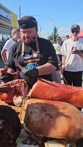 A man in a black t-shirt and apron with "HRENNOV BBQ" on it, wearing a black knit cap and sunglasses, is standing at a wooden table outdoors. He is holding a large knife in his right hand and a piece of cooked meat in his left. Several large cuts of cooked meat, including what appears to be a whole pig's head, are on the table in front of him. In the background, there are other people, some watching the man. A yellow building with "HAVASS" written on it is visible in the far background. The scene appears to be an outdoor event or market.