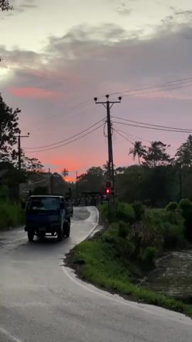 The video shows a rural street scene at sunset. An Isuzu truck passes the camera on a wet road. A railroad crossing with flashing red lights is visible in the distance, framed by trees. The sky displays a mix of grey, pink, and orange hues as the sun sets. Utility poles line the road, and lush green vegetation covers the roadside. A person in a red shirt walks past the camera after the truck.