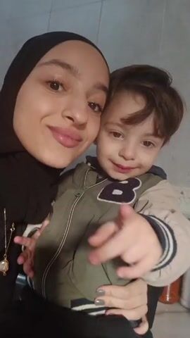 A woman wearing a black hijab is holding a young child in her arms. The woman is smiling and looking at the camera. The child is looking to the side. They appear to be in a kitchen, as there are various jars and containers on the counter behind them and tiled walls in the background. The woman seems to be taking a selfie or a short video with the child. There is lively music in the background.