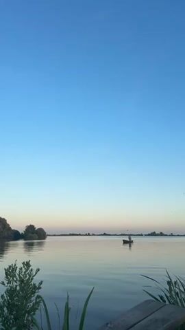 The video shows a calm lake at sunset. The sky is a clear blue, transitioning to a pale pink near the horizon. On the water, a small dark boat with a single person is visible in the middle ground, with a person standing in the boat. The shoreline is lined with trees and vegetation, which are reflected in the still water. In the foreground, a wooden bench and some green plants are visible. The overall scene is peaceful and serene, capturing the beauty of nature at dusk.