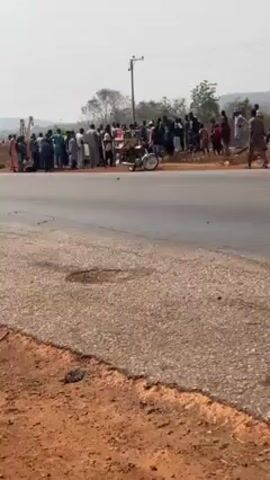 The video shows a group of people gathered on the side of a road, near what appears to be an accident scene. There is a three-wheeled motorcycle (tricycle) on the road near the group. The people are of varying ages and are dressed in a mix of traditional and casual clothing. The road appears to be paved but with some visible wear. The surrounding environment includes dry, reddish soil and some sparse vegetation. In the distance, there are hills and a power line. The sky is overcast, suggesting a hazy or slightly dull day. The people are congregated, presumably observing the aftermath of the incident, though their specific actions or interactions are not clearly discernable in the static frame.