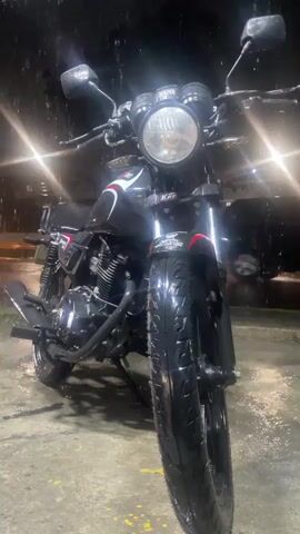 A black motorcycle with red and white accents is parked on a wet surface. Rain is falling, creating streaks in the air and making the ground shiny. The motorcycle has a prominent round headlight and two mirrors. The background is dark with blurry lights that suggest a nighttime urban environment. The front tire of the motorcycle is facing the camera, and its tread is visible and wet.