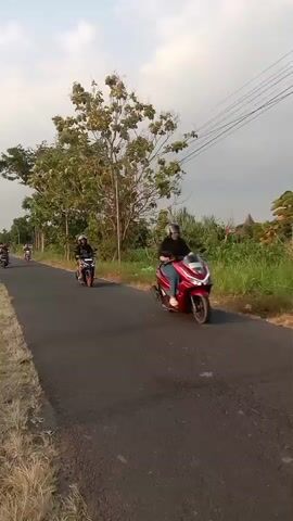 A person is riding a motorcycle on a paved road. The person is wearing a helmet and dark clothing. The motorcycle is black and has orange wheels. The road is bordered by grass and trees. There are power lines overhead. The sky is cloudy. The time is approximately dusk.