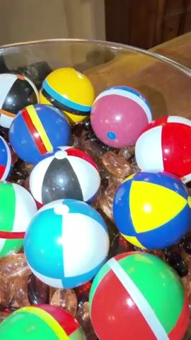 The video displays a close-up, rotating view of a clear glass bowl filled with small, colorful balls. The balls are primarily beach balls with various color combinations, including red, white, blue, yellow, green, and black. Some balls feature a smooth, spherical surface, while others appear to have a slightly textured finish. The balls are resting on a bed of small, amber-colored, translucent glass beads or gems. The background is blurred, suggesting a shallow depth of field, but hints at a wooden surface and dimly lit interior. The camera movement is slow and deliberate, allowing for a comprehensive examination of the contents of the bowl. No people or actions are visible, only the static arrangement of the balls and decorative elements.