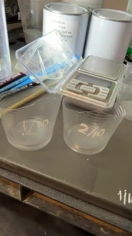 The video shows a close-up view of a surface, likely a workbench or table, with various items laid out for what appears to be a measuring or mixing process. In the background, two white metal cans are visible, one with a label in Hebrew. Next to them is a clear plastic container holding some white powdery substance. A digital scale is placed on a clear plastic lid. In the foreground, several clear plastic cups are arranged. These cups are marked with handwritten numbers, suggesting they are for different measurements or trials. Some cups contain white powder. The surface itself has handwritten markings, including numbers and what appear to be abbreviations. A small pile of white powder is scattered on the surface. A pink object, possibly a stick or applicator, is also present. The overall impression is one of a meticulous, scientific or artistic process involving precise measurements of powders.