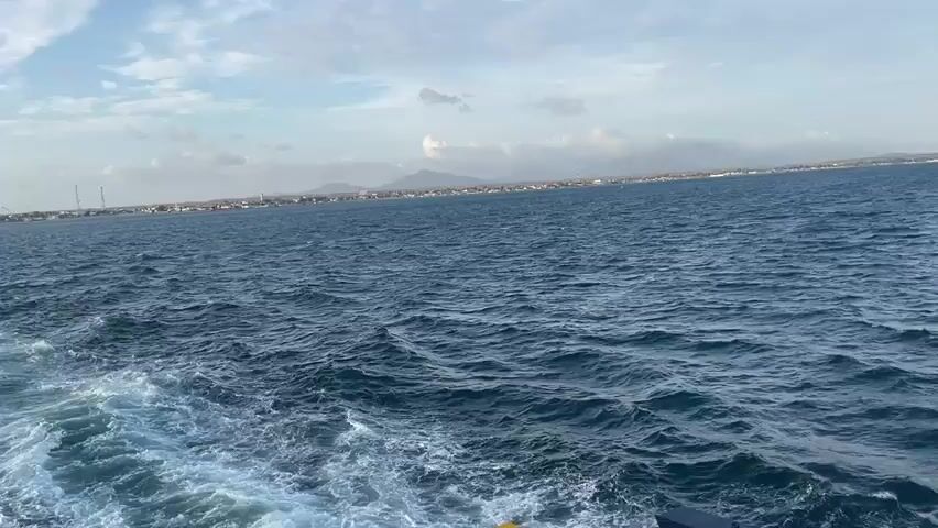 The video shows a view from the deck of a moving vessel, looking out at a body of water. The water is choppy with white wakes trailing behind the vessel, indicating motion. In the distance, a coastline is visible with buildings and what appear to be mountains or hills in the background. The sky is partly cloudy with a mix of blue sky and white clouds. Part of the vessel's deck is visible in the foreground on the left and right sides, including some railing and a piece of equipment. The overall scene suggests a journey across the sea.
