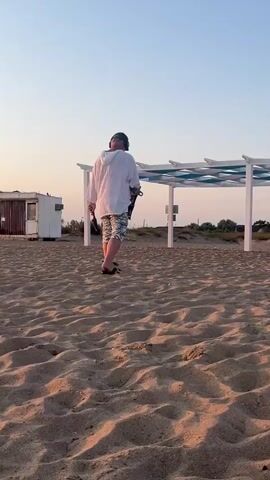A man is metal detecting on a sandy beach near a white structure with a blue and white patterned roof. He is wearing a white hoodie, camouflage shorts, a baseball cap, and headphones. He is holding a metal detector in his right hand and a small shovel in his left hand. The sun is setting, casting long shadows on the sand. There are other people in the background, some walking and some sitting.