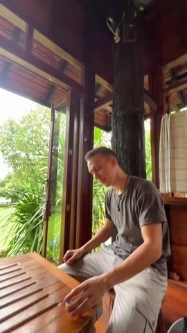 A man in a grey t-shirt and grey pants sits at a wooden table with a slatted surface. He holds a long, flat, wooden object in his hands and appears to be cleaning or smoothing it with a dark object. In the background, through an open doorway, a lush green lawn stretches out to a body of water, possibly a rice paddy, with trees and clouds visible in the distance.