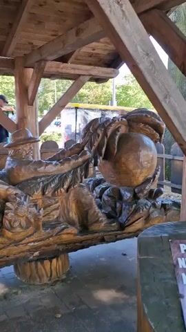 The video shows a wooden sculpture located under a wooden shelter. The sculpture appears to be a scene with several carved figures and objects. In the foreground, a large carved log forms the base of the sculpture, with carved figures of people and animals, including what looks like a man with a hat and possibly chickens. In the background, there is a train-like structure with people standing near it. There are also cars visible in the distance. The ground in front of the sculpture is wet, creating reflections of the carving. A person in a red jacket walks past the sculpture, and a young girl in a pink jacket is also seen looking at the sculpture. The environment appears to be outdoors, possibly a park or a recreational area.