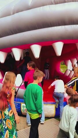 A close-up vertical shot of children playing inside a vibrant pink inflatable shark bouncy castle, its white teeth on full display. A young boy in a black cap and red shirt is in the foreground, his face partially obscured, looking down at the activity. Other children are scattered throughout the inflatable structure, some standing, some sitting, all seemingly enjoying themselves. 

The child closest to the camera on the right side of the frame has long, reddish-blonde hair and is wearing a colorful patterned dress. To their left, another girl in a bright pink top and light blue jeans stands beside a boy in a green long-sleeved shirt and black pants. More children are visible further inside the shark's mouth, with one boy in a white shirt and black shorts, and another in a black and white striped shirt. 

The bouncy castle is set up outdoors on a paved area next to a patch of green grass. The weather appears fair, suggesting an outdoor event or a sunny day. In the background, beyond the inflatable structure, some faint text is visible, possibly identifying the location as "Ulbroka, Latvia." The atmosphere is lively and joyful, with the children actively engaged in play.