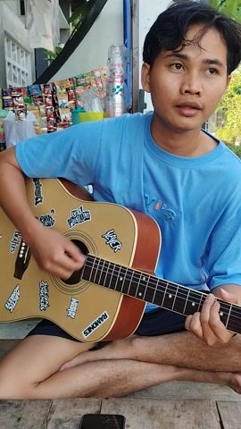 A young male is seated outdoors, cross-legged on a wooden surface, playing an acoustic guitar. He is wearing a blue t-shirt and dark shorts. The guitar has several stickers on its body, including ones that appear to say "SEX PISTOLS," "RAMONES," and "JIMI HENDRIX." His right hand strums the strings while his left hand is positioned on the fretboard.

Behind him, a white wall is visible, with a rack of various packaged goods, possibly snacks or toiletries, displayed against it. The area appears to be an open-air setting, with some greenery and trees in the background, suggesting it might be daytime. Sunlight illuminates the scene. A smartphone is visible on the wooden surface in front of him. The overall mood seems relaxed and casual. The location is identified as Pakuncen, Indonesia.