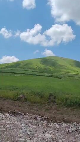 A low-angle, static shot captures a rolling green hill under a bright blue sky dotted with fluffy white clouds. The foreground is a dirt path littered with small stones and gravel. The hill is covered in lush green grass with visible terraced lines indicating cultivation or erosion control. No people or animals are visible in the frame. The overall impression is a serene, natural landscape.