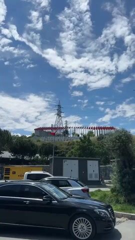 The camera faces a green hill in a city. A tall, metal, electrical transmission tower stands in front of the hill. Behind the hill, is a gray building. The hill has a sign that says, "15 TEMMUZ GAZIOSMANPAŞA" in large, white letters.  Rows of Turkish flags wave in the wind. The sky is bright blue with scattered white clouds. In the foreground are several parked cars, and a gray, electrical power unit sits on the side of the road. Electrical lines are strung across the top of the scene.