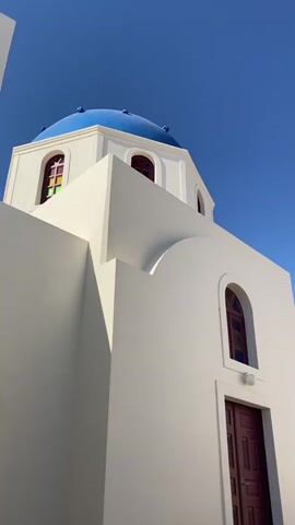 The video shows a low-angle, upward shot of a white building with a blue dome, set against a clear blue sky. The building appears to be a church or a similar structure, with arched windows and a wooden door. The sunlight casts sharp shadows, indicating a bright day. In the distance, a faint crescent moon is visible in the sky. The camera pans slightly to the right, revealing more of the building's architecture and its surroundings, which include other white buildings and hints of vegetation. A security camera is visible mounted on one of the walls. Further to the right, two people are walking away from the camera, one with a white shirt and green patterned shorts, and another partially visible person. The scene suggests a warm, sunny day, possibly in a Mediterranean or similar climate, given the architectural style.