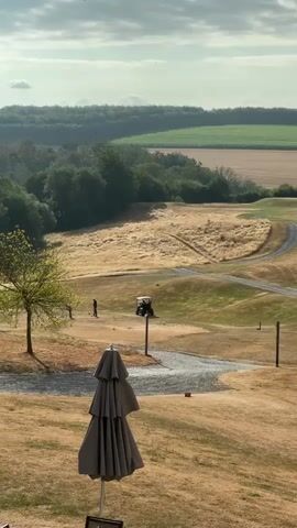The video shows a golf course on a sunny day. In the foreground, a closed umbrella is visible. Further back, two people are on the golf course, one of whom appears to be preparing to hit a golf ball. A golf cart is parked nearby. The landscape is rolling hills covered in dry grass and trees, with a line of darker green trees in the distance. The sky is partly cloudy.