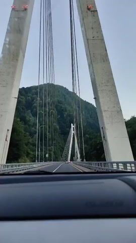 This video shows a first-person perspective, likely from inside a vehicle, as it travels across a suspension bridge. The bridge is characterized by tall, white concrete towers and numerous cables that fan out to support the roadway. The road itself is paved and has clear lane markings, including a white line on the left and a yellow line on the right. Guardrails line both sides of the bridge.

In the distance, a smaller, white A-frame structure, also part of the bridge's support system, is visible. Beyond the bridge, a lush, green mountainside rises steeply, densely covered with trees. The sky is a pale blue with some haziness, suggesting either early morning or late afternoon, or possibly overcast conditions. A small, faint object, possibly the moon or a distant airplane, is visible in the upper left quadrant of the sky.

Streetlights are positioned along the bridge. There are a few other vehicles on the bridge, appearing as distant specks. The overall scene is one of natural beauty with impressive man-made infrastructure. The "SRS AIRBAG" text is visible on the dashboard in the foreground, confirming the viewpoint is from inside a car. The location is identified as Kepsha, Russia. The activity is driving across the bridge. No people are clearly discernible. The mood is serene and observational, focused on the scenic drive.