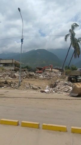 This is a video of a street scene, likely following a disaster. The camera is mounted vertically, giving a landscape view. In the foreground, there is a paved road with yellow curb markings. To the left of the road, there is a large pile of debris, consisting of broken concrete, twisted metal, and rubble. A tall telecommunications tower stands behind the debris, with a large billboard attached to it. In the background, there are mountains and a cloudy sky. A few palm trees are visible amidst the debris. There are no people visible in the video. The overall impression is one of destruction and aftermath.