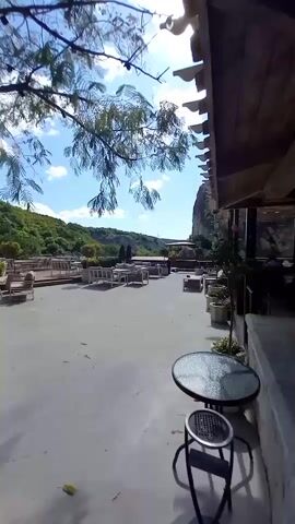 The video shows an outdoor patio area with tables and chairs, likely a restaurant or cafe, set against a backdrop of lush green hills under a bright blue sky with scattered clouds. In the foreground, a round glass-topped table with a dark metal stool sits on a concrete floor. A tree with green foliage and a single red rose is visible to the left of the frame. The patio extends into the background, featuring multiple rows of tables and seating arrangements. The sun is shining, casting shadows and creating a warm, inviting atmosphere. The overall impression is of a peaceful and scenic outdoor dining or relaxation space.