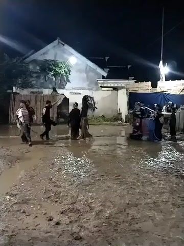 At night, a group of people, primarily men, are gathered in a very muddy, unpaved outdoor area. The ground is thoroughly saturated and churned into thick mud, suggesting recent rain. In the background, a simple house with a gabled roof is visible, illuminated by artificial lights, along with some wooden structures and what appears to be a blue tarp or tent.

The people are engaged in a lively activity. Some individuals are holding whips and moving dynamically, while others walk or interact. At one point, a man in a striped shirt falls or is pulled to the ground in the mud. Another man, wearing a dark shirt, drags him briefly before he stands up. A tall, dark, effigy-like figure, possibly an animal prop related to a performance, stands in the middle ground. The scene appears to be a cultural performance or celebration taking place in Jenggrik, Indonesia, given the setting and the dynamic, whip-wielding actions. No specific text is clearly visible.