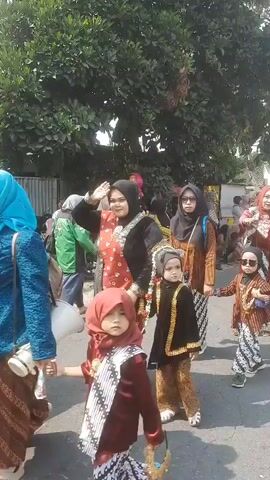 The video shows a procession of people, primarily women and children, walking down a paved street lined with trees and buildings. Most of the participants are wearing traditional Indonesian clothing, including batik patterns and headscarves. Several children are dressed in formal attire, with some wearing crowns or ornate headwear. The atmosphere appears festive, with people smiling and interacting with each other. The camera is handheld and follows the procession from the side. The lighting suggests it is daytime, with sunlight filtering through the trees.