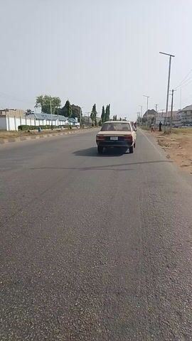 The video shows a daytime scene on a paved road, likely in an urban or semi-urban area. The perspective is from a vehicle, possibly a yellow commercial mini-van (identifiable by "TOYOTA" written on its rear). Ahead of this vehicle is a beige-colored van, emitting some exhaust, followed by another yellow vehicle further down the road. Several pedestrians are visible walking or standing on the unpaved shoulder to the left of the road. The road surface shows cracks. Utility poles line the side of the road, and sparse greenery and distant buildings are visible. The sky is bright and clear, indicating sunny weather. The scene depicts everyday traffic and pedestrian movement at Kaduna Junction, Nigeria.