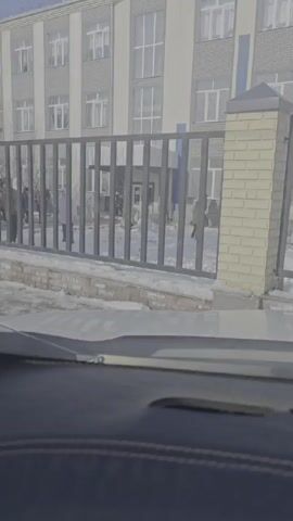 The video, shot from inside a vehicle, shows a school building with multiple floors, framed by a dark metal fence. Patches of snow line the base of the fence. A group of people, mostly clad in winter clothing, stands in front of the school's entrance, visible through the fence. The sky is bright, suggesting a sunny day.