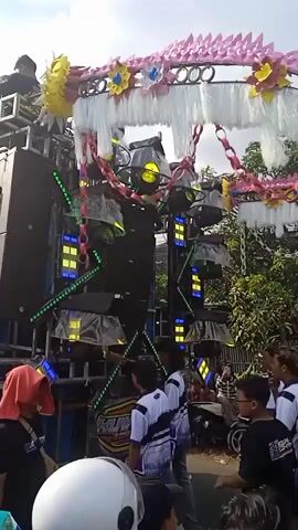 The video shows a large, mobile sound system at what appears to be an outdoor event. The system is decorated with colored lights and streamers. Several people are gathered around, some wearing matching white and blue shirts. There are trees in the background. The sound system is emitting smoke.