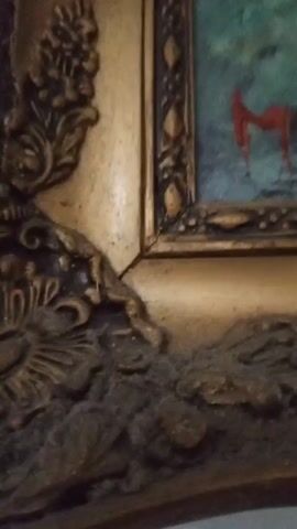 The video shows a close-up of a painting's frame and a portion of the painting itself. The frame is ornate and gold-colored, with intricate designs. The painting, visible above and to the right of the frame, appears to have a blurred, abstract style with muted greens, blues, and a stroke of red that resembles a signature. The signature seems to read 'MUKLIS' in red ink. The background is a plain white surface. The camera seems to be moving slightly during the recording, and there is audible speech in the background.