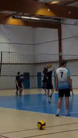 The video shows a volleyball game taking place in an indoor sports hall. Several young men wearing sports jerseys are on a blue volleyball court. In the foreground, a person wearing a white and blue jersey with the number 7 on the back is standing and watching the action. Further away, another person wearing a dark jersey with the number 3 is spiking a volleyball over the net. The volleyball is yellow and blue. The net is standard volleyball net. In the background, there are banners and advertisements on the wall. The hall has wooden beams supporting the roof.