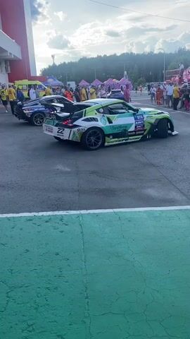 Two drift cars are parked on an asphalt surface. The car on the left is blue and white with various sponsor logos. The car on the right is white, green, and black with sponsor logos and the number 22 visible. There are people standing around the cars, some in yellow shirts. In the background, there are tents, trees, and a building with a red and white facade. The sky is cloudy. The cars are stationary.