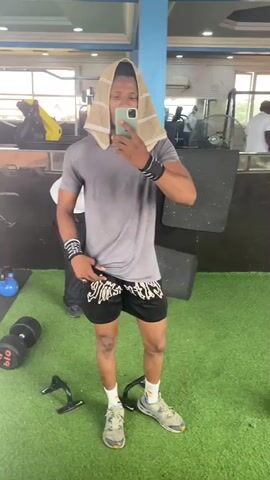 A man wearing a grey t-shirt, black shorts, wrist wraps, and athletic shoes stands in a gym. He has a towel draped over his head and is holding a phone to take a mirror selfie. He flexes his bicep as he takes the photo. Other gym equipment, including dumbbells and workout machines, are visible in the background. Another person is partially visible behind him.