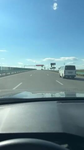 The video shows a view from the dashboard of a car driving on a multi-lane highway. The sky is clear blue with a few white clouds. The highway has a concrete barrier on the left and a guardrail on the right. There are white lane markings on the road. In the distance, there are large overhead signs. A white van is driving in the right lane ahead of the car. The car appears to be moving at a steady speed. The dashboard of the car is visible in the foreground.