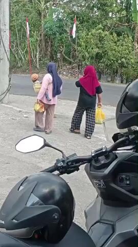 Two women are walking away from the camera on a paved path. The woman on the left is wearing a light pink shirt, a dark blue hijab, and light brown pants. She is carrying a baby on her back in a sling and a yellow basket in her left hand. The woman on the right is wearing a black shirt, a dark red hijab, and checkered pants. She is carrying a yellow plastic bag in her right hand. Both women are walking towards a road with trees and foliage in the background. Several Indonesian flags are visible on poles along the road. In the foreground, a motorcycle and a black helmet are parked.