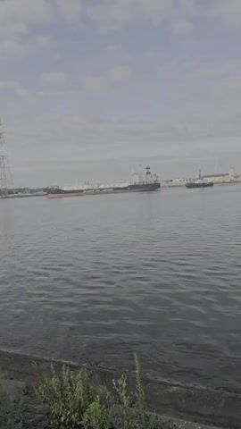 A wide shot captures a view of a body of water with a large ship in the distance. The water is choppy, with small waves. On the left side of the frame, a tall metal tower is visible, possibly a power line pylon. The ship is long and appears to be a cargo or tanker vessel, painted white with a black superstructure and a red hull line. It is positioned in the middle of the water, moving away from the viewer. Further in the distance, on the right side of the frame, another, smaller vessel can be seen. The sky is overcast with scattered clouds. The foreground shows the edge of a bank with some sparse green vegetation growing. The overall lighting suggests it is daytime.