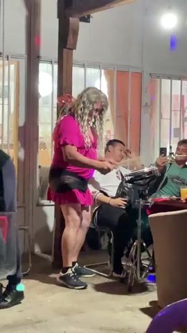 Outside on a patio, three men and one person dressed in drag are visible. The person in drag, who appears male, wears a pink top and matching pink mini skirt with a black fanny pack. The person has a blonde wig and is wearing sunglasses. They are holding drumsticks and appear to be playing a small set of drums. One man is sitting behind the drums, not interacting. A second man sits at a table with a glass of orange juice, holding a pipe to his lips, seemingly about to smoke it. A third man stands in the foreground on the left. The patio has a concrete floor and is surrounded by windows. Festive lighting is present.