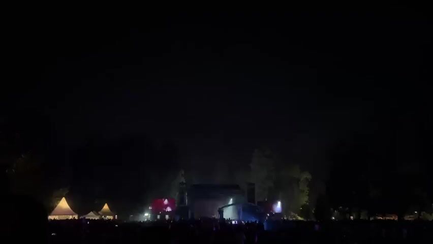 This is a nighttime video of an outdoor concert. A large crowd of people is gathered in front of a stage. The stage is illuminated by bright lights, and there are large screens on either side of it. There are also tents set up in the background. The sky is dark, and there are trees visible behind the stage. The overall atmosphere appears to be festive and energetic.