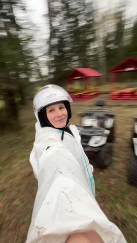 The video captures a person, likely a woman, filming herself in a selfie style in an outdoor, wooded environment. She is wearing a white full-body protective suit with light blue accents, a white full-face helmet featuring a clear visor and the visible text 'K.Y.T.' on the side, and a black balaclava. She smiles broadly, conveying a sense of happiness and excitement.

Behind her, several dark-colored All-Terrain Vehicles (ATVs) are parked on a ground covered with dry leaves and patchy grass. At least one other person, also wearing a helmet and dark attire, is visible near the ATVs. The background features a dense line of trees, suggesting the edge of a forest, and a distinctive red gazebo-like structure further back.

The camera dynamically spins around the main subject, creating a playful and energetic visual effect. The diffused lighting indicates an overcast daytime. The presence of protective gear and ATVs strongly suggests participation in an ATV riding or outdoor adventure activity in a natural, possibly recreational, area like Zubovo, Russia.