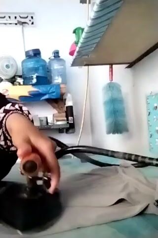The video shows a person ironing a piece of light-colored fabric on an ironing board. They are holding an iron in their right hand and moving it across the fabric. Steam is visible emanating from the iron. In the background, there are shelves with various items, including water bottles, a fan, and cleaning supplies. A blue feather duster hangs from a shelf above the ironing board. The person is wearing dark clothing.