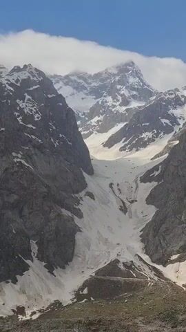 The video shows a mountainous landscape with snow-covered peaks under a partly cloudy sky. The mountains are primarily gray with patches of snow clinging to their sides. A large snow-filled gully runs down between the mountains towards a rocky base. The video appears to be taken from a moving vehicle.