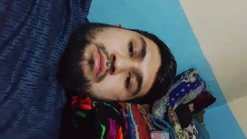 The video shows a man lying down on a bed with his head resting on some colorful fabric. He is looking at the camera with a slightly tired expression. He has dark hair and a beard. He is wearing a patterned shirt. The background consists of a wall painted in two shades of blue and off-white.