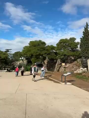 The video shows a stone walkway with several people strolling on it. To the right of the walkway is a rocky hill with trees. A signpost with a map or information panel stands near the edge of the walkway. At the top of the rocky hill, some people are sitting. The sky is blue with white clouds. The scene appears to be a park or historical site with people exploring the area.