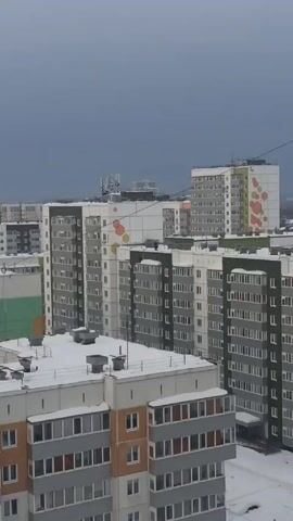 The video shows a view of several multi-story apartment buildings on an overcast day with some snow on the rooftops. The camera zooms in on one building where two people are visible on the roof near a ladder. One person is standing while the other appears to be seated or crouching down near the ladder.