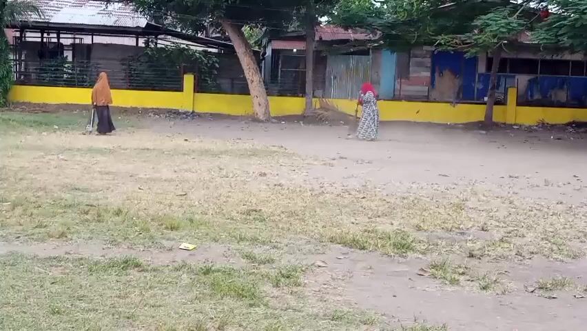 The video shows a dirt and grass area with two women in the background. The woman on the left is wearing a brown hijab and a dark dress, and is holding a broom. She appears to be sweeping. The woman on the right is wearing a red hijab and a patterned dress. There are buildings and trees in the background, and a yellow wall runs along the back of the area.