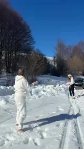 Two women are playing in the snow on a sunny day. The woman on the left is wearing a white sweatsuit, and the woman on the right is wearing black leggings and a white top and hat. They are throwing snowballs at each other in a snowy area with trees in the background and tire tracks in the snow. There are also a couple of parked cars visible in the distance.